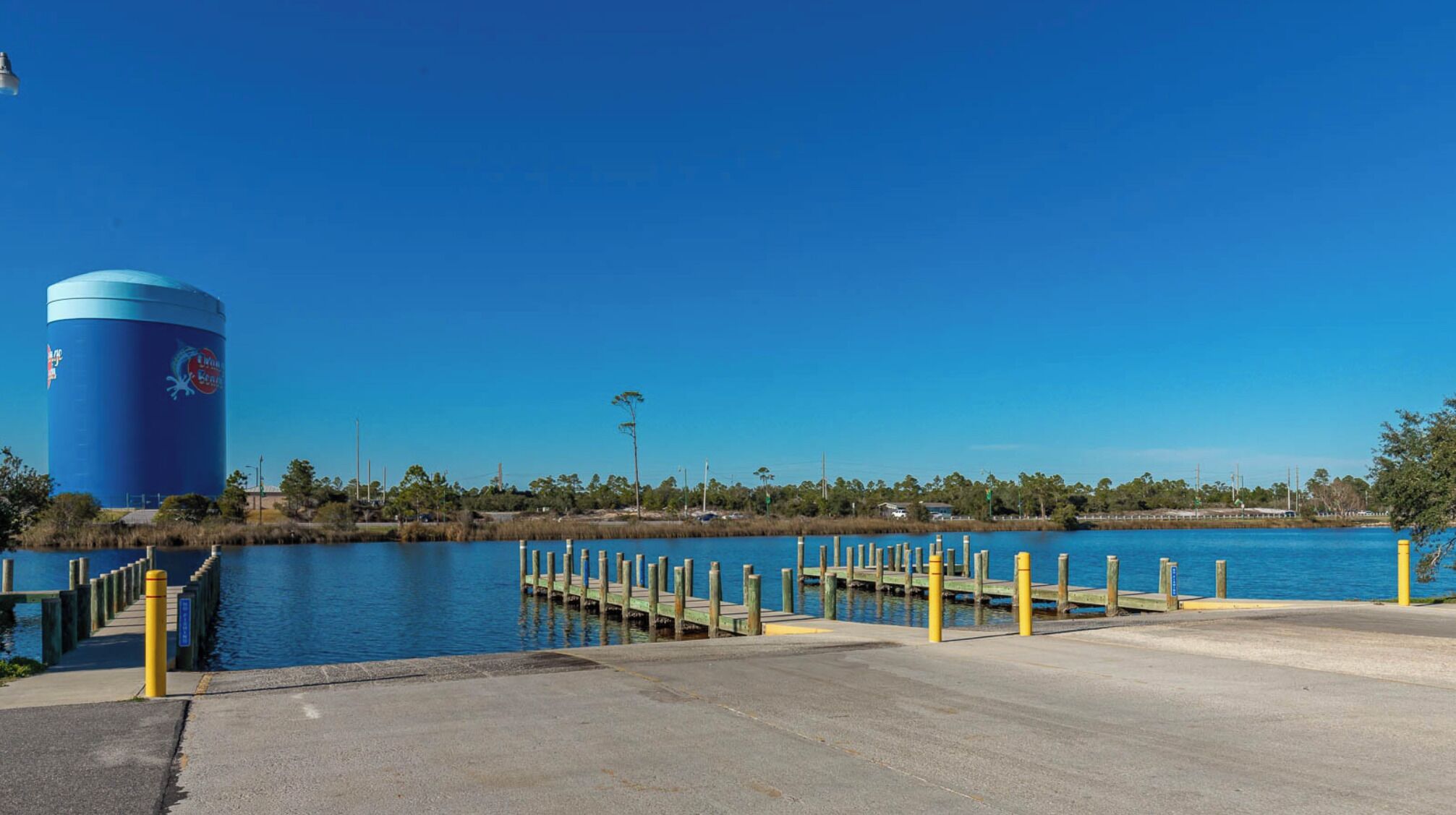 A waterfront area with a large blue cylindrical structure, a concrete dock with yellow posts, and a clear blue sky.