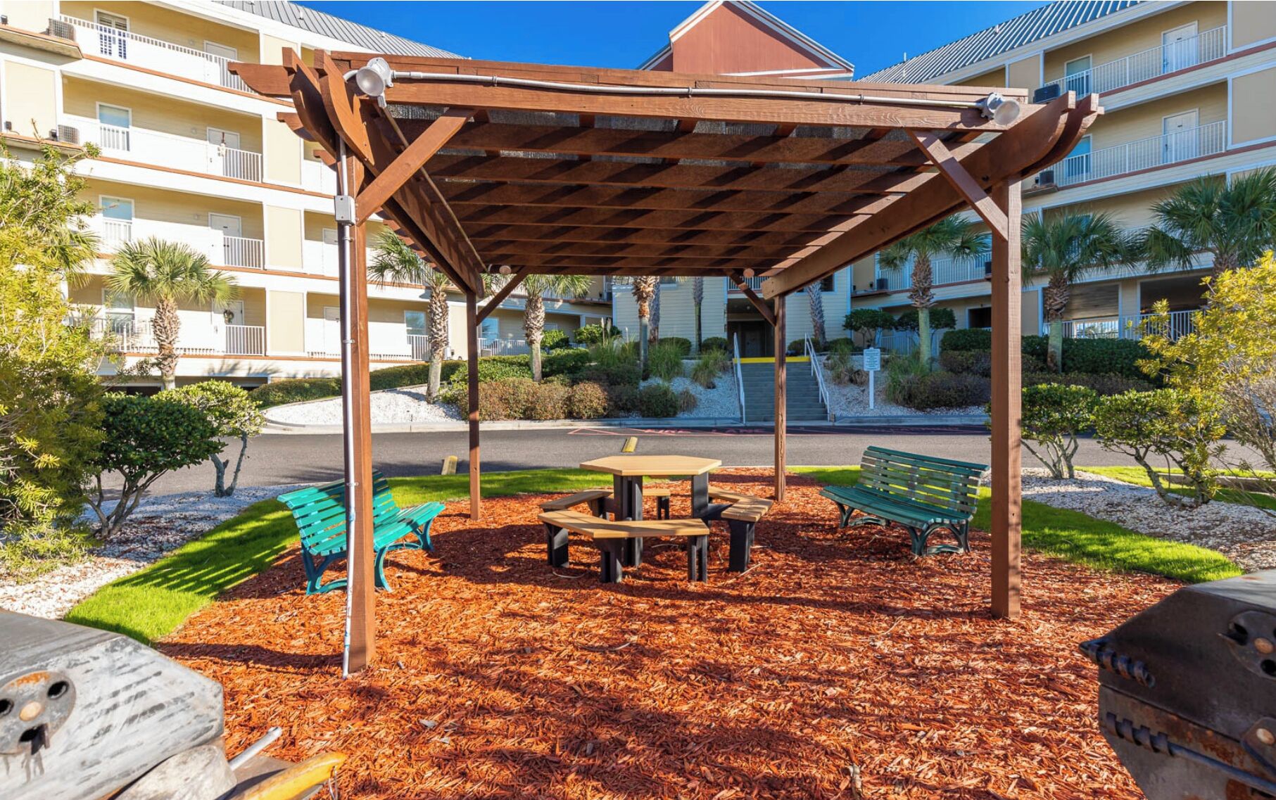A covered outdoor seating area with a table and benches, surrounded by mulch and small trees, in front of a multi-story building with balconies.