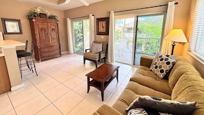 A living room with a sofa, coffee table, dining table, and chairs. There is a cabinet, a ceiling fan, and a window with curtains.