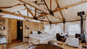 Ein gemütlicher, rustikaler Zeltraum mit Holzboden, einem runden Holztisch, einem Bett mit weißer Bettwäsche, einem Kamin und einer Sitzecke mit Sessel und Sofa.