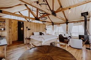Een gezellige, rustieke tentkamer met een houten vloer, een ronde houten tafel, een bed met wit beddengoed, een open haard en een zithoek met een stoel en een bank.