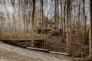 A rustic cabin with a wooden deck and stairs leading up to the main structure, surrounded by a forested area.