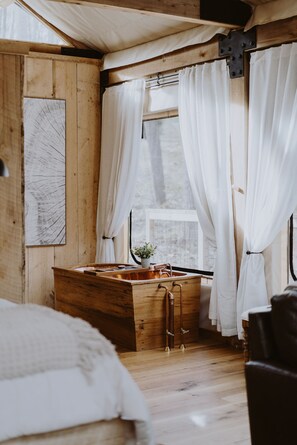 A wooden cabin interior with a wooden bed, a wooden dresser, and a window with white curtains.