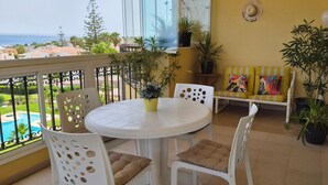 Un balcón con una mesa redonda y sillas, plantas en macetas y vistas a una piscina y edificios.