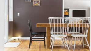 Uma área de refeições com uma mesa de madeira e cadeiras brancas, uma cadeira preta e dois quadros nas paredes.