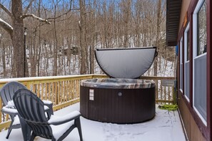 A hot tub on a wooden deck with a chair, surrounded by snow-covered trees.