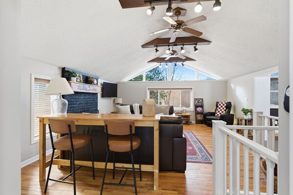 A spacious living area with a high ceiling, a wooden dining table with bar stools, a black sofa, and a television.
