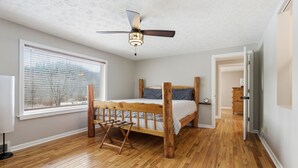 Quarto com cama de madeira, ventoinha no teto, janela com persianas e porta que dá para outro cômodo.