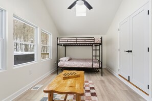 A room with a bunk bed, a wooden table with a chessboard, a window with a view of trees, and a white door.