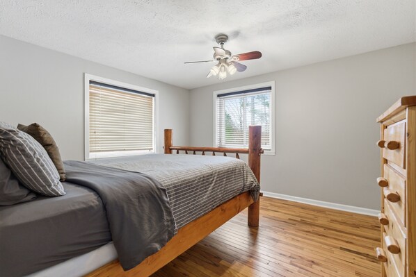 A bedroom with a wooden bed, two windows with blinds, a ceiling fan, and a wooden dresser.