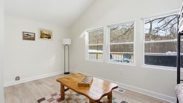 A room with a wooden table, a checkered rug, two windows with a view of trees, a floor lamp, and two wall-mounted pictures.