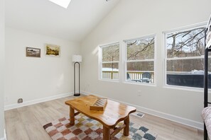 A room with a wooden table, a checkered rug, two windows with a view of trees, a floor lamp, and two wall-mounted pictures.