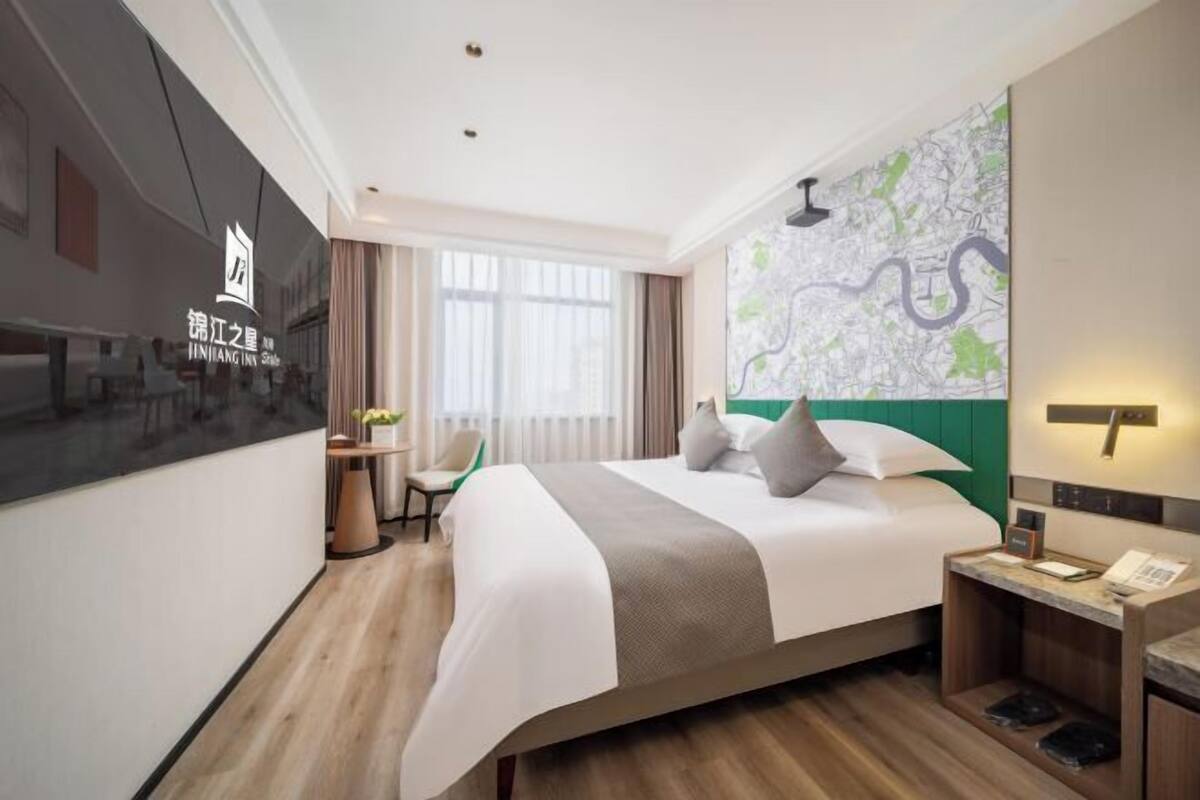 A hotel room with a bed, a desk, a chair, a map on the wall, and a TV on the left.