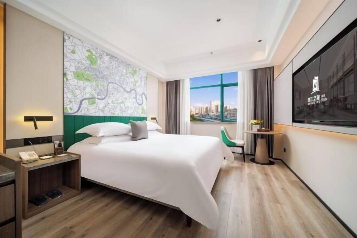 A hotel room with a large bed, a desk with a phone, a map on the wall, and a view of the city through a window.