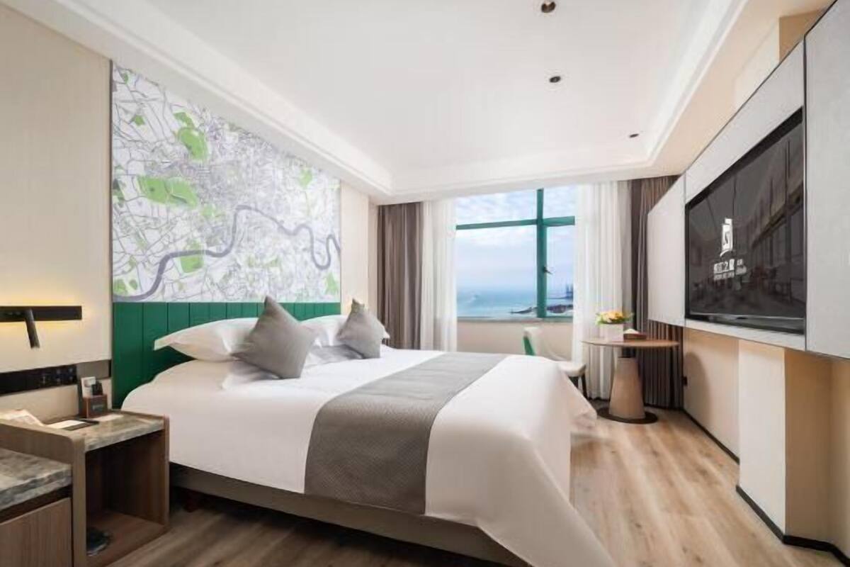 A modern hotel room with a large bed, a map artwork on the wall, a TV, and a view of the sea through the window.