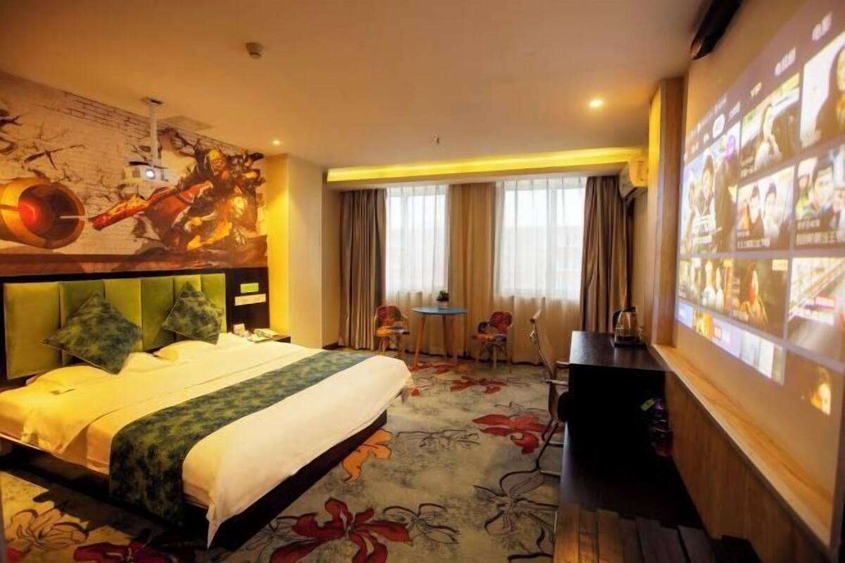 A hotel room with a bed, a desk, a chair, a small table, and a large screen displaying images.
