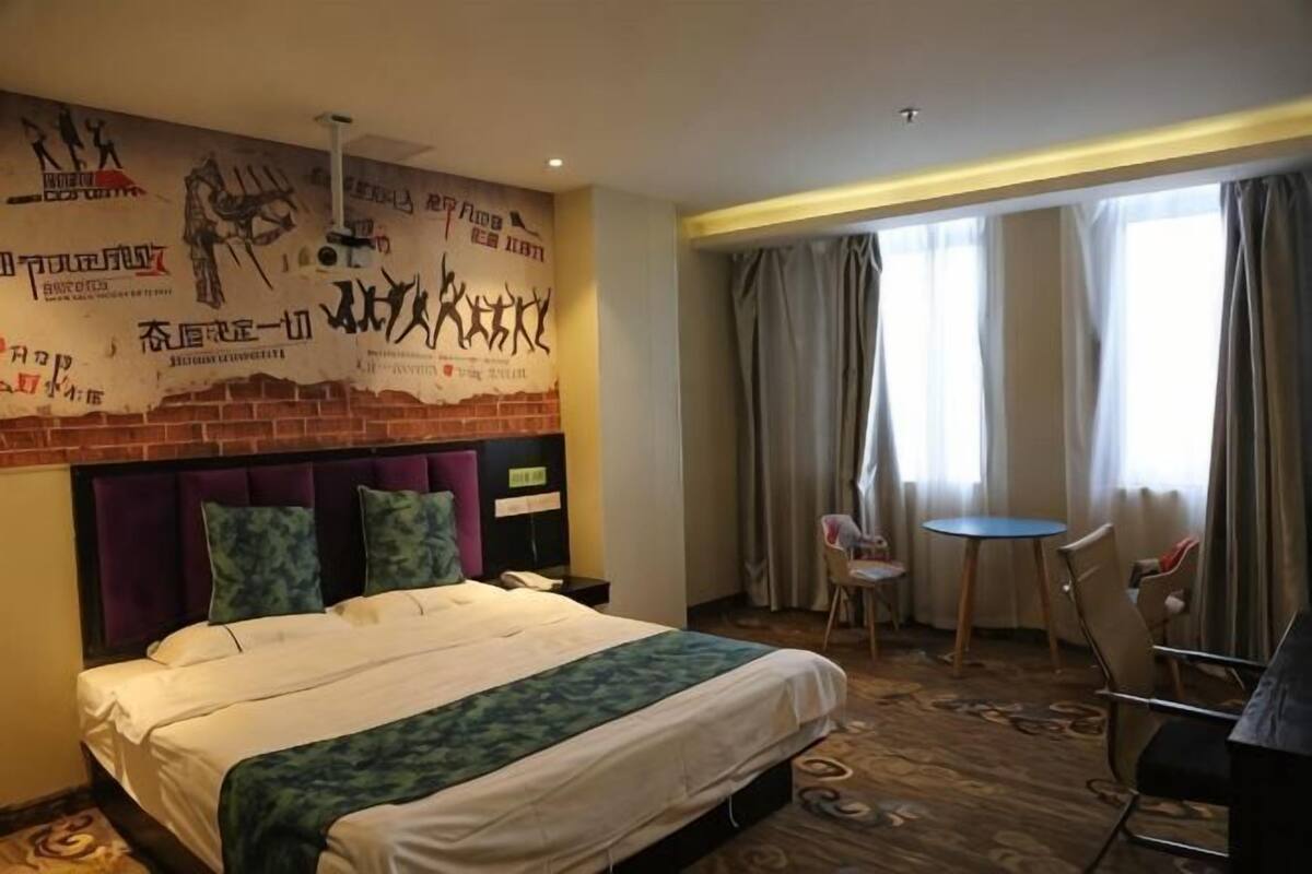 A hotel room with a bed, a small table, two chairs, a wall with artwork, and a window with curtains.