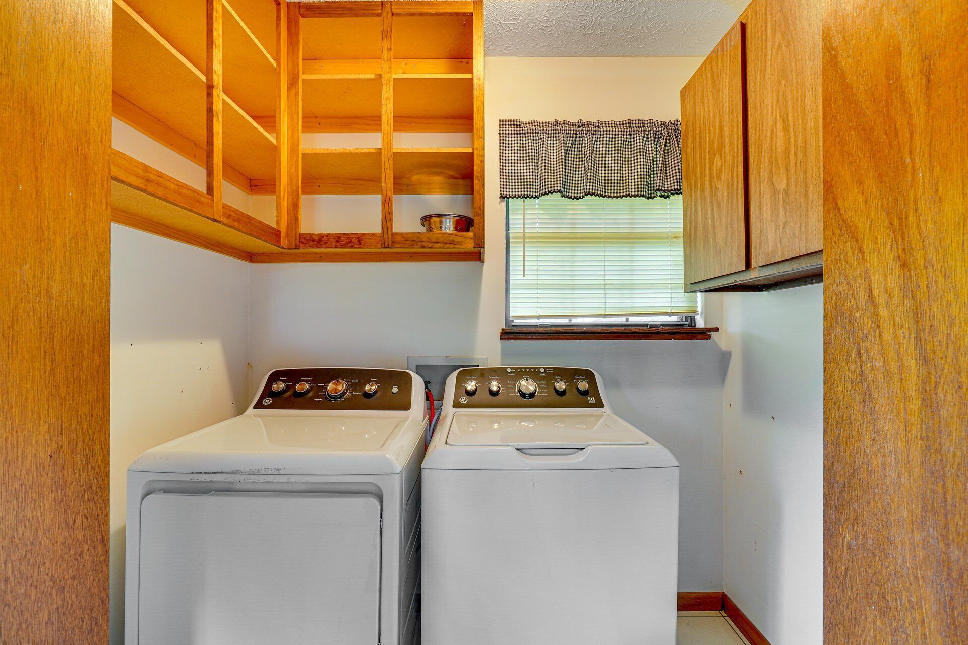A laundry room with two stacked washers and dryers, wooden cabinets, a window with blinds, and a checkered valance.