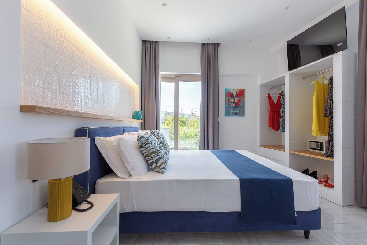 Ein modernes Hotelzimmer mit einem großen Bett, einem Kleiderschrank mit Kleidung, einem Nachttisch mit Lampe und Blick ins Freie.