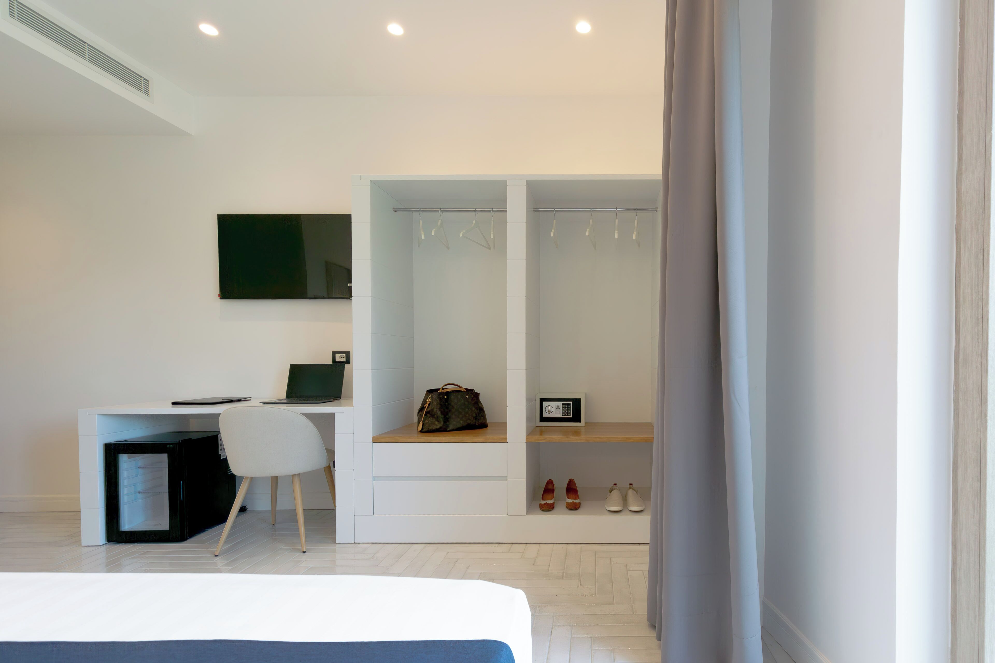 A modern hotel room with a bed, a desk with a laptop, a chair, a wardrobe, and a shoe rack.