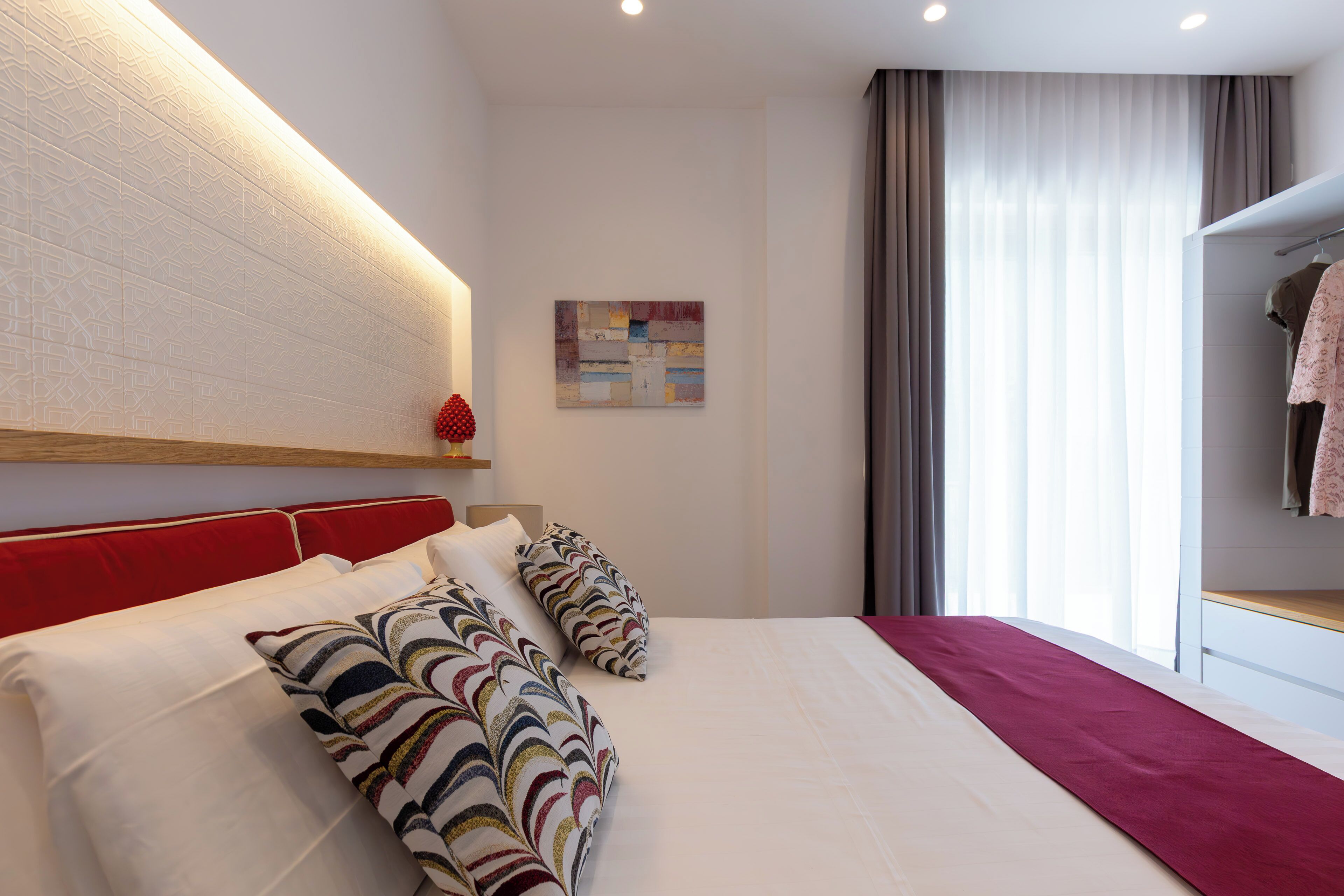 Une chambre d’hôtel avec un grand lit, une tête de lit rouge, une étagère fixée au mur et une fenêtre avec des rideaux.