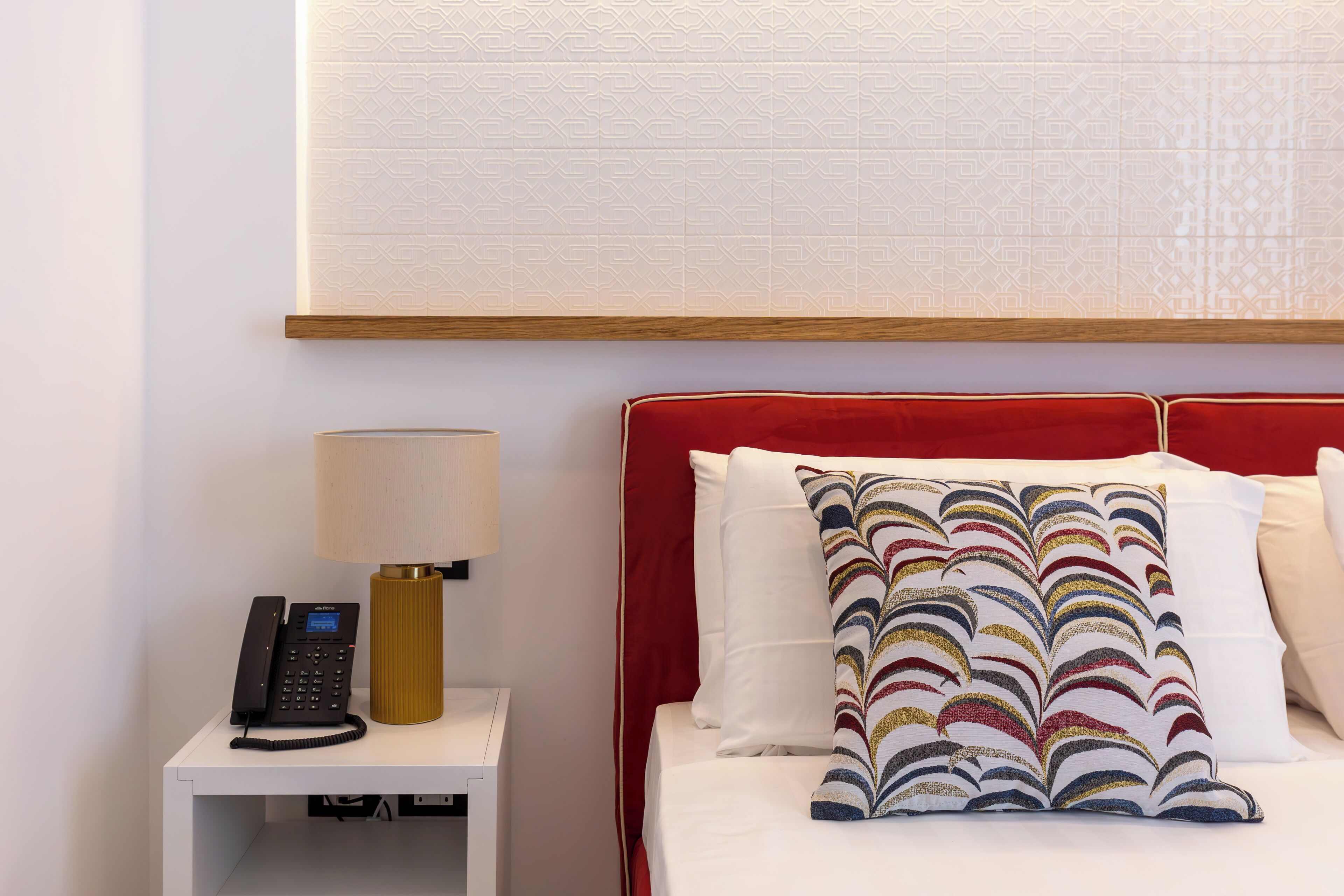 A hotel room with a bed, a nightstand with a phone and lamp, and a patterned wall.
