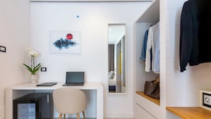Ein modernes Hotelzimmer mit weißem Schreibtisch, einem Laptop, einem kleinen Kühlschrank, einem Stuhl, einem Spiegel und einem Kleiderständer mit Kleidung.