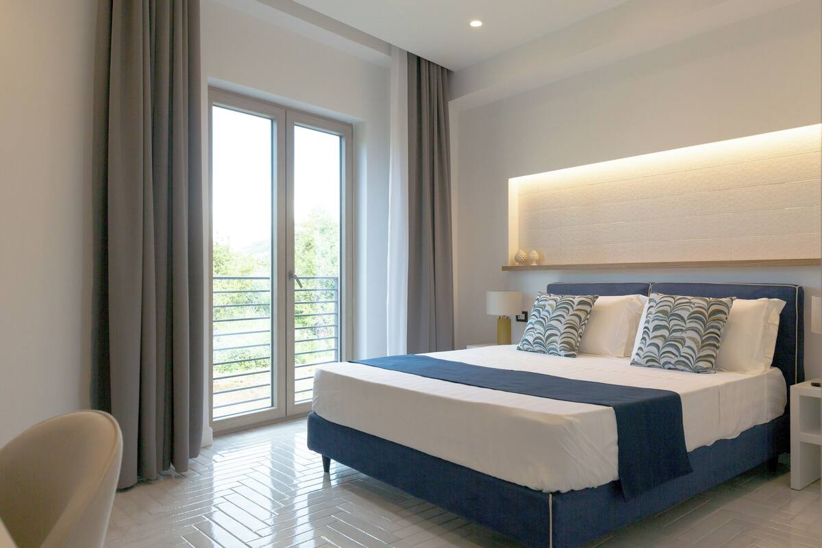 Ein modernes Hotelzimmer mit einem großen Bett, einem Nachttisch, einem Sessel und Blick auf einen Balkon.