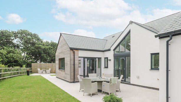 Et moderne hus med terrasse, spisebord og stole, et mindre skur og et stort vindue.