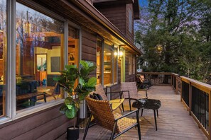 Une terrasse en bois, avec des chaises et une table en osier, donnant sur un bâtiment aux grandes fenêtres et offrant une vue sur les arbres.
