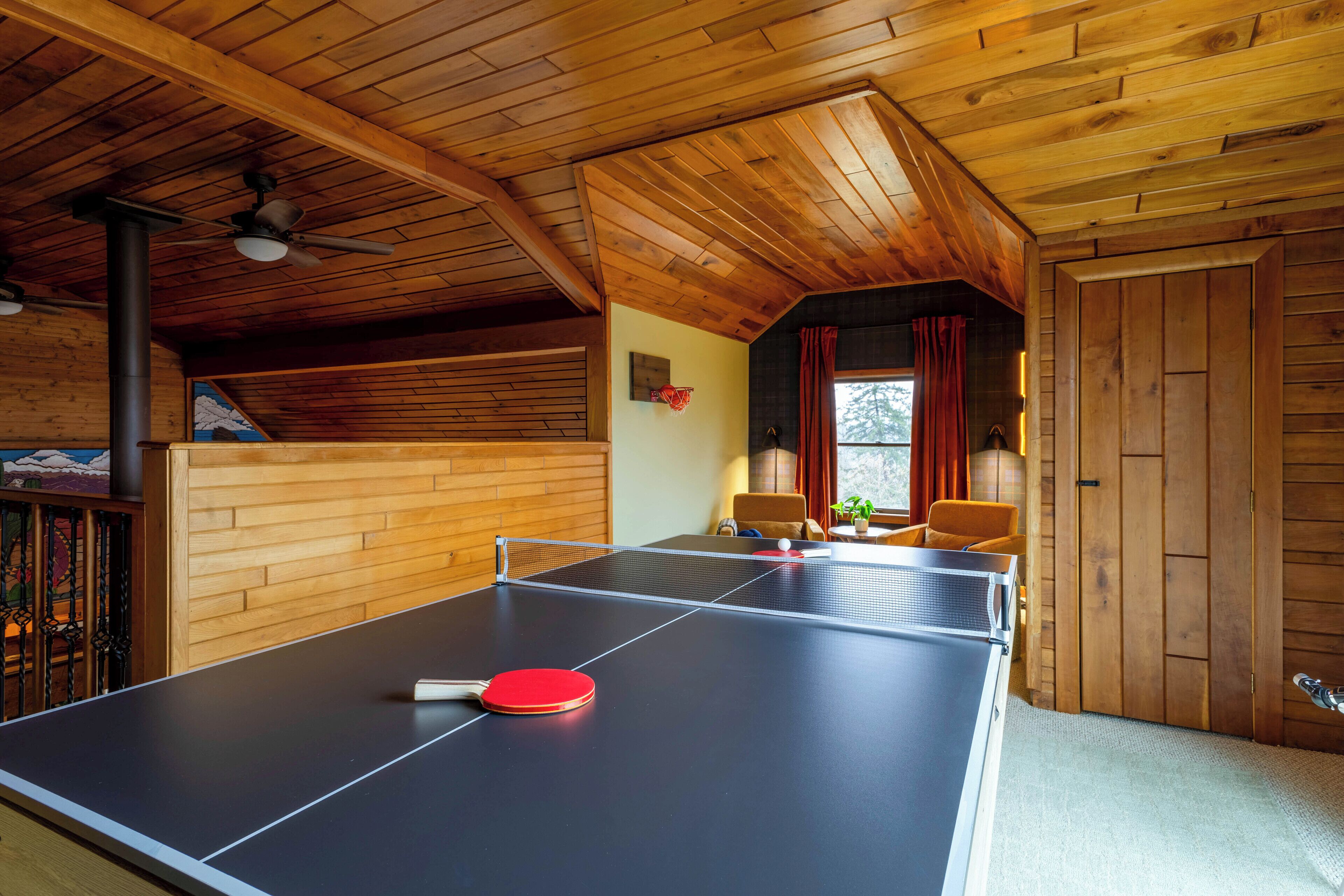 Une table de ping-pong se trouve dans une pièce en bois, dotée d’un ventilateur de plafond, d’une porte et d’une fenêtre avec des rideaux.