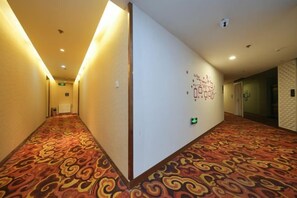 A hotel corridor with patterned carpet, a white wall with a decorative design, and a green emergency exit sign. - Ward Aladdin Hotel (Changjiang East Road Huangdao Golden Beach Branch) (Qingdao)