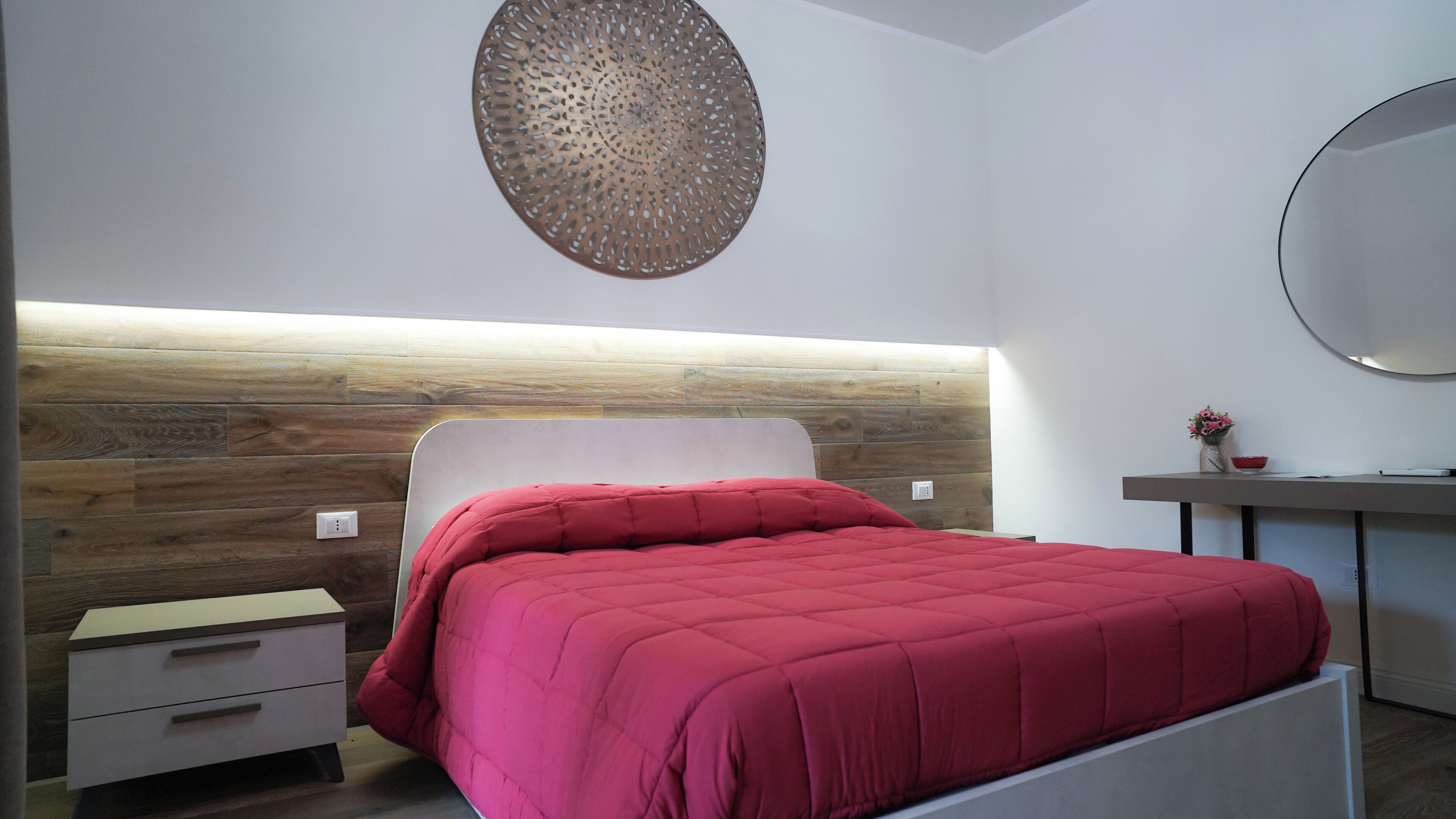 A modern bedroom with a wooden headboard, a red bedspread, a bedside table with a lamp, and a round mirror on the wall.