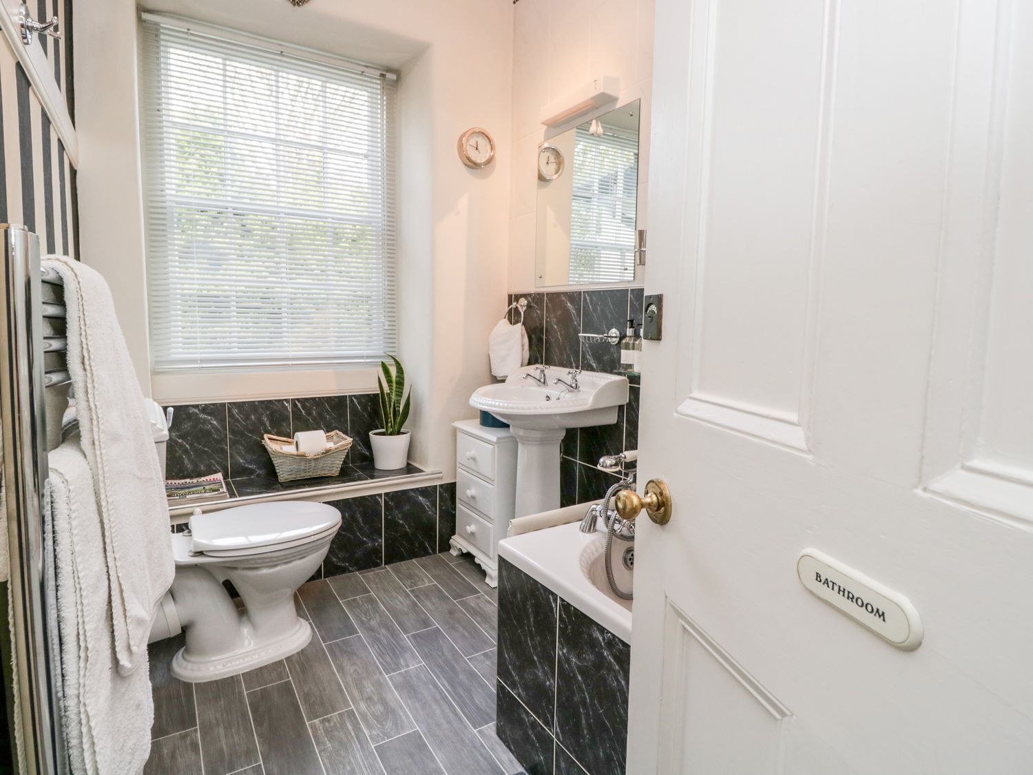 A bathroom with a toilet, sink, and mirror. There is a window with blinds, a towel rack, and a plant.