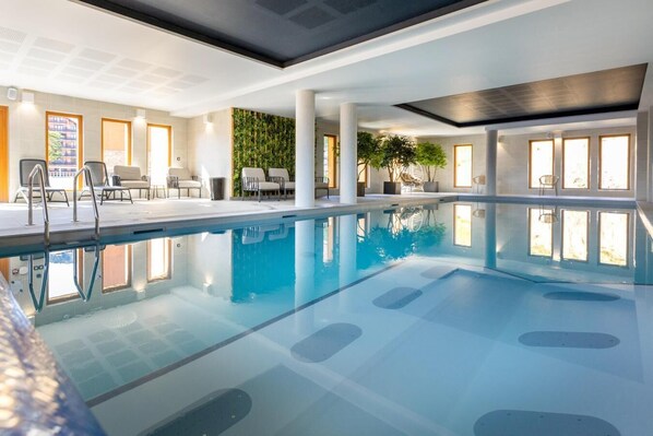 A modern indoor swimming pool with clear water, surrounded by white walls and large windows.
