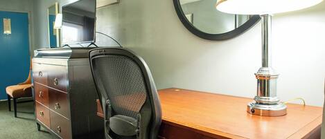 A hotel room with a wooden desk, a black office chair, a lamp, a television, and a dresser.