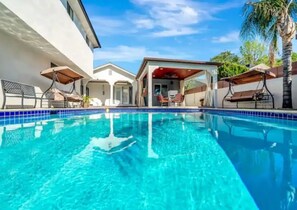 Zwembad met helderblauw water, omringd door een wit gebouw en een overdekt terras met stoelen en parasols.