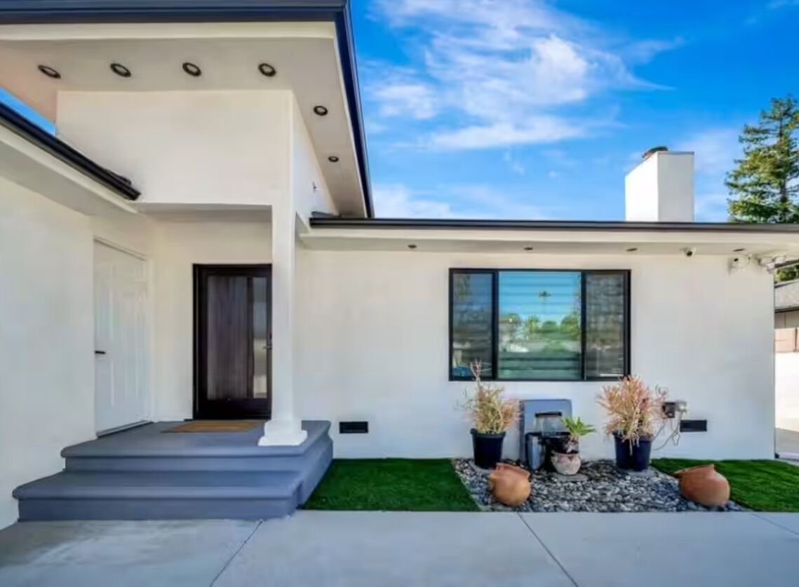 Una casa moderna con techo plano, un porche con escalones y un pequeño jardín con plantas en macetas.