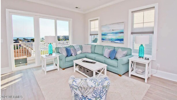 A spacious living room with a light blue sofa, a patterned armchair, and a coffee table. There are two white side tables with lamps and decorative items. A large window offers a view of the outside. - Posh, New, Ocean View Townhome in gorgeous Emerald Isle Perfect for families (Emerald Isle)