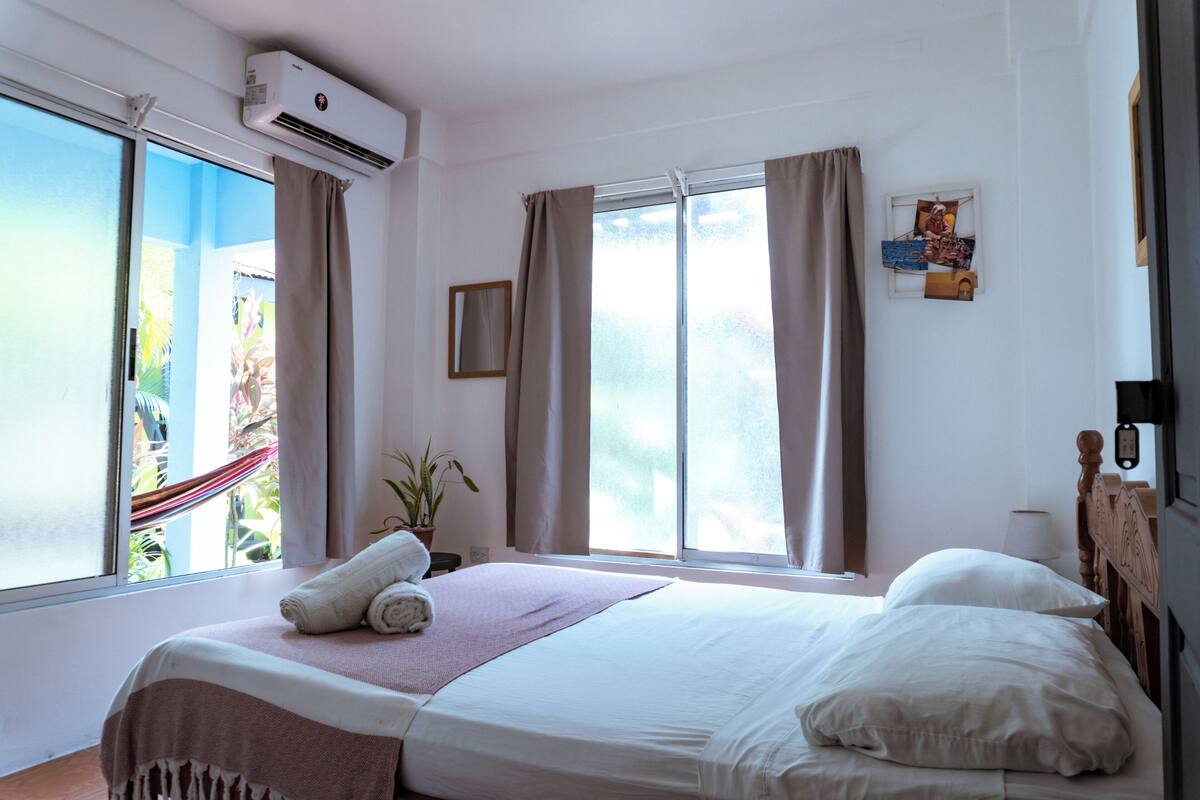 Quarto de hotel com cama, unidade de ar condicionado, janelas amplas, cortinas, um vaso de planta e vista para o exterior.