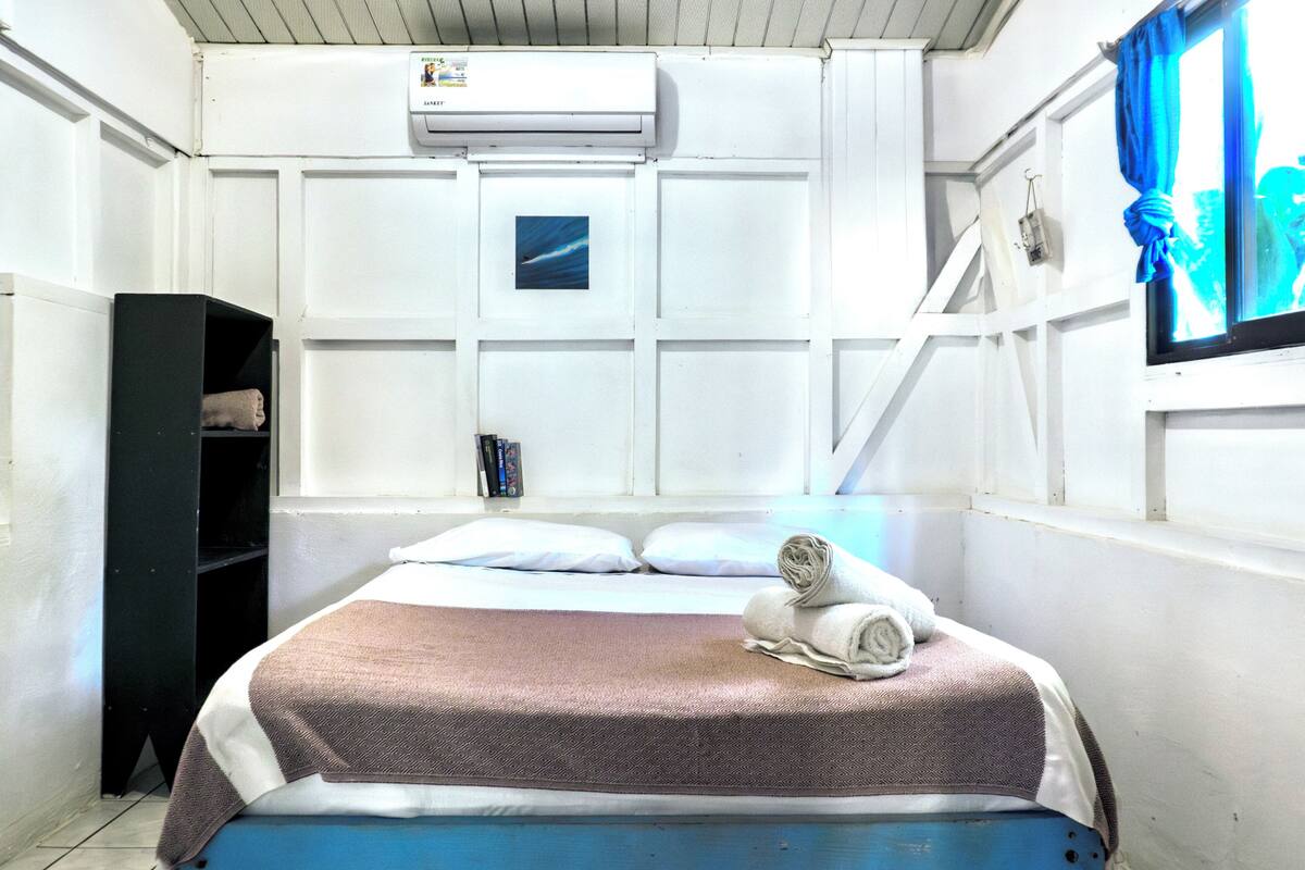 Quarto com cama, prateleira, ar condicionado e janela com cortina.