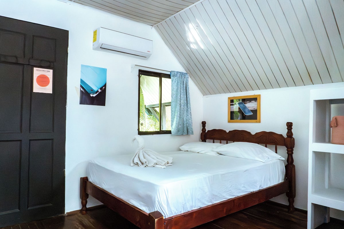 Quarto com cama de madeira, roupa de cama branca, janela com vista, ar condicionado e porta.