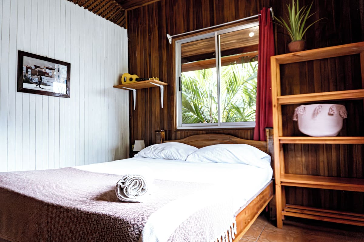 Uma cama com estrutura de madeira, roupa de cama branca, uma toalha enrolada e uma prateleira com um vaso de planta.