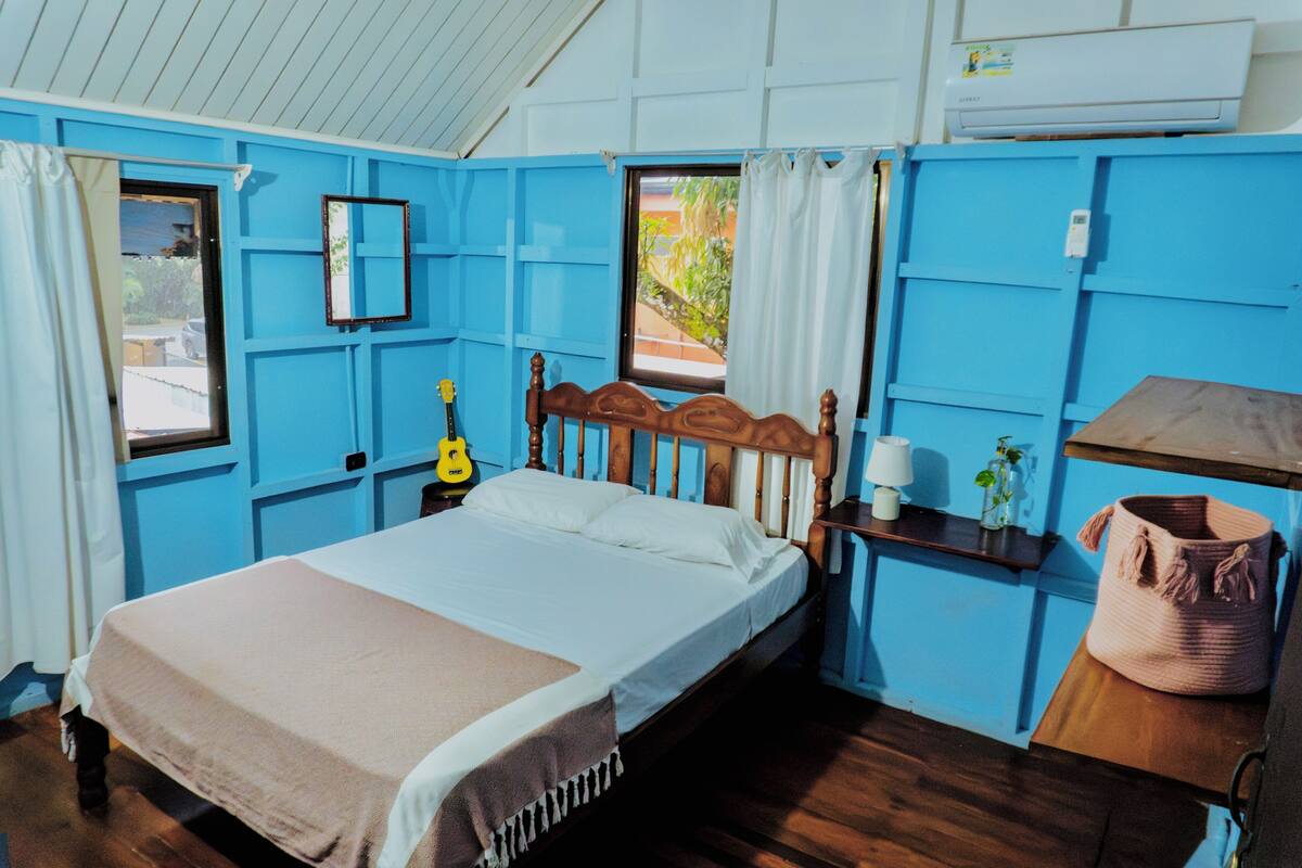 Um quarto com uma cama de madeira, um violão, uma mesa de cabeceira, um abajur e uma cesta grande de fibra natural.