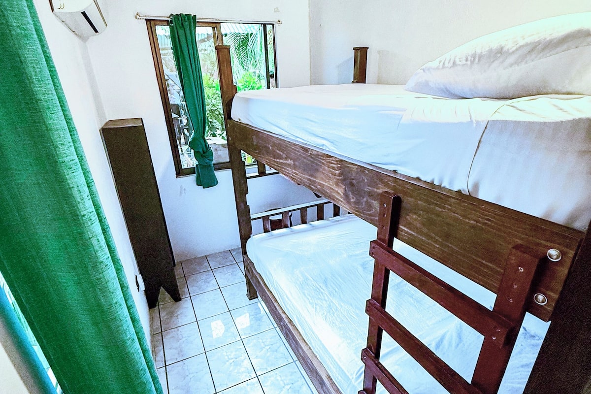 Quarto pequeno com uma cama individual em beliche, chão de azulejos, janela com cortinas e uma cortina verde parcialmente visível.