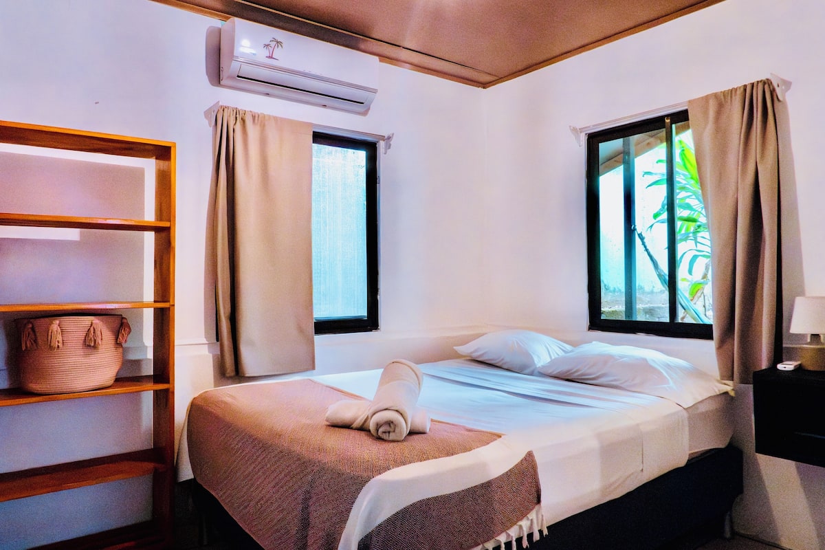Uma cama de solteiro com uma toalha enrolada, uma prateleira com um cesto e uma janela com cortinas.