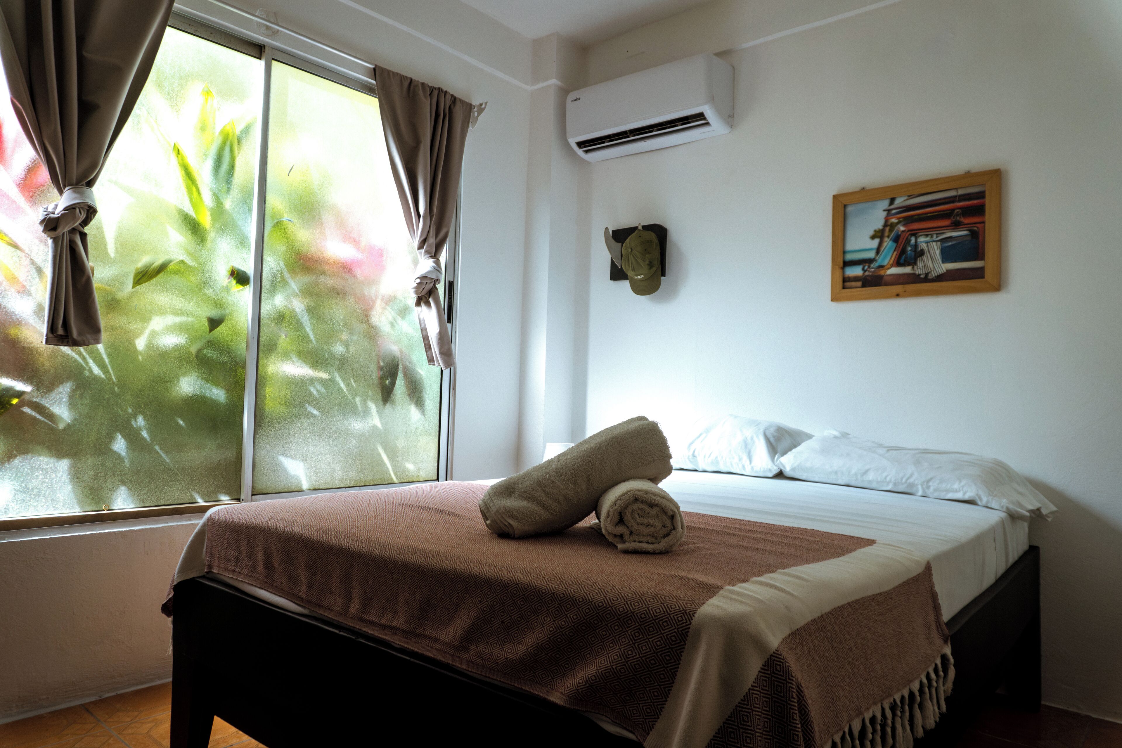 Ein Hotelzimmer mit Bett, einem fensterbepflanzten Fenster, einer Klimaanlage und einem gerahmten Bild an der Wand.