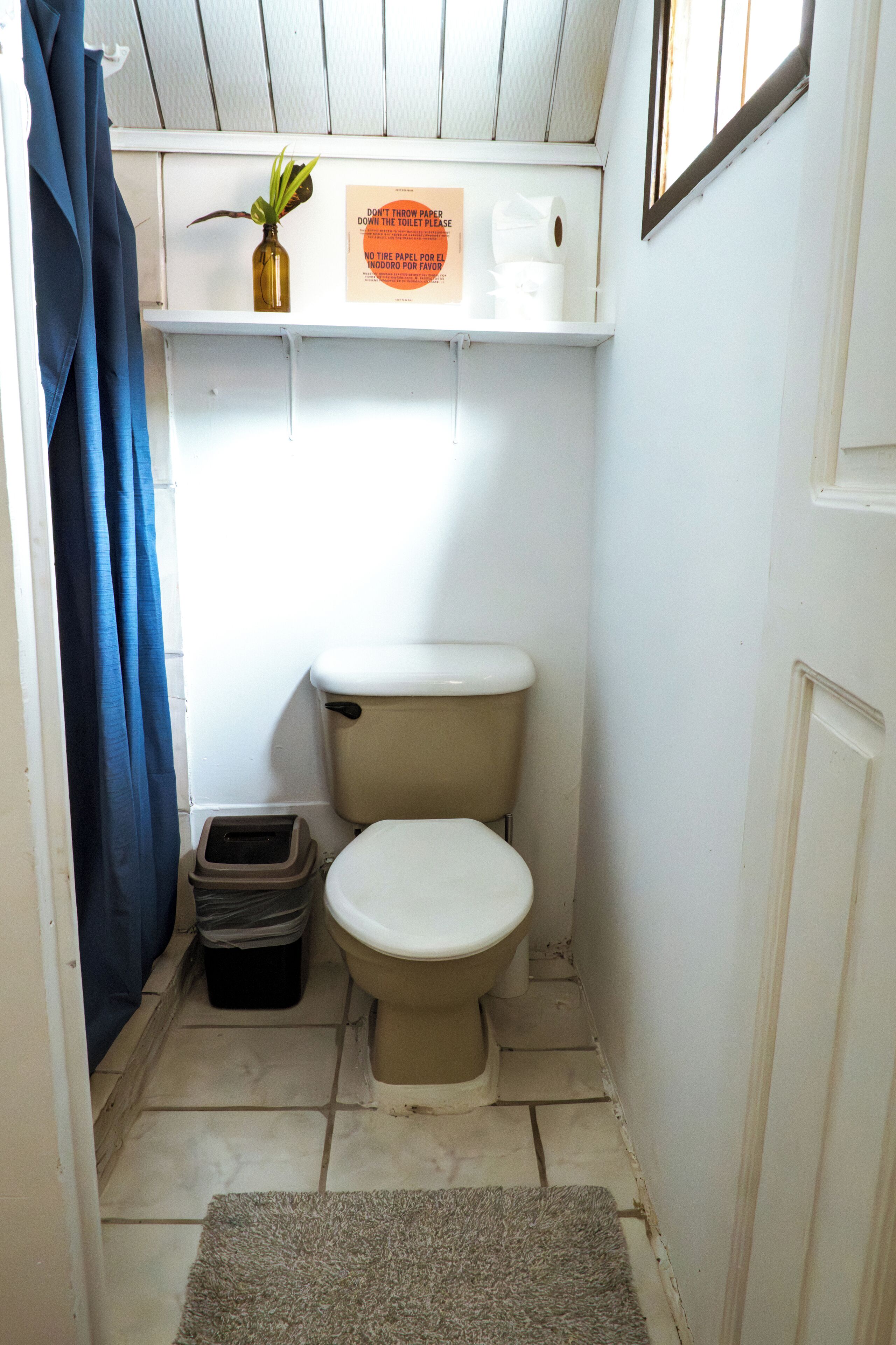 A small, compact bathroom with a toilet, a trash bin, a shelf with a plant and a sign, and a blue curtain.