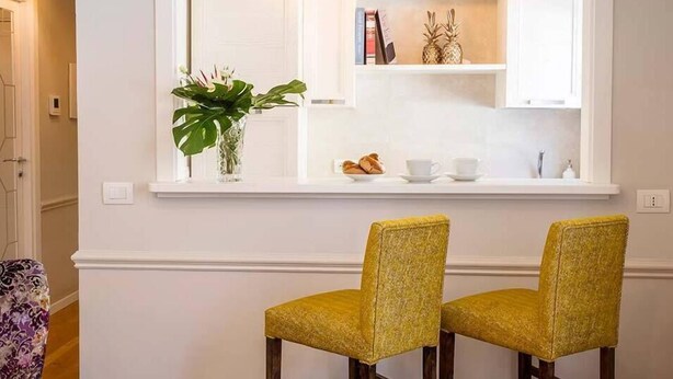 Een moderne keuken met witte kasten, een inbouwplank met boeken en decoratieve objecten, en twee gele stoelen ervoor.