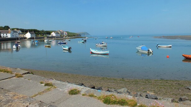 A harbor with various boats anchored in calm waters, a rocky shoreline, and buildings in the background. - Castell Y Garn (Newport)